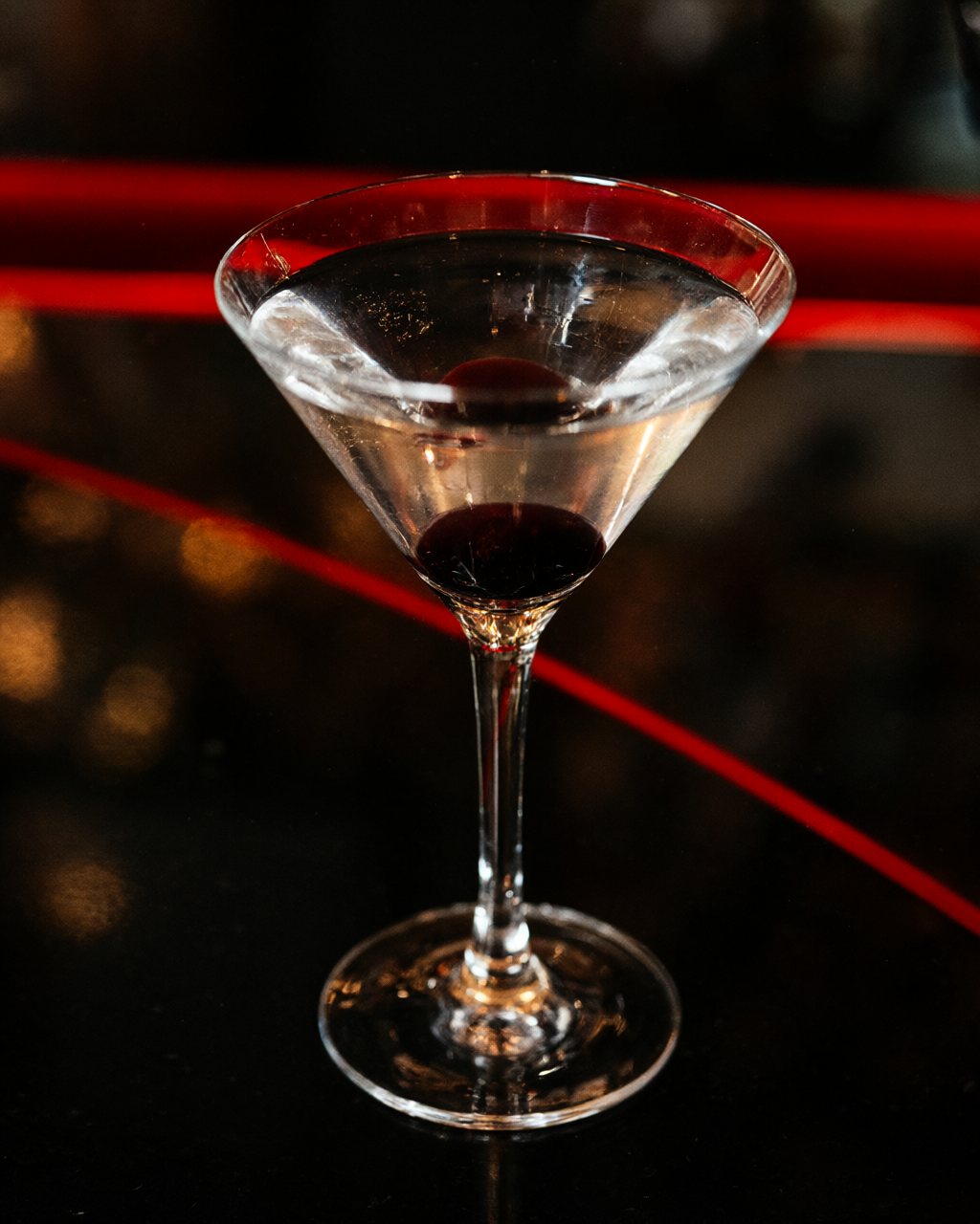 A martini with a single dark cherry on a black bar, lit in red