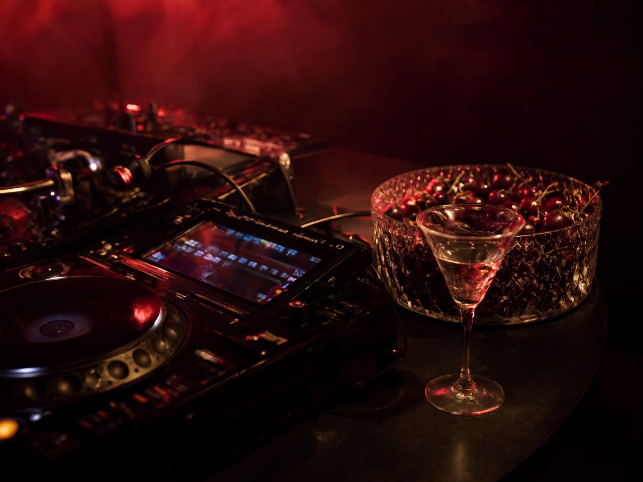 A DJ booth in red light with a bowl of cherries beside the decks